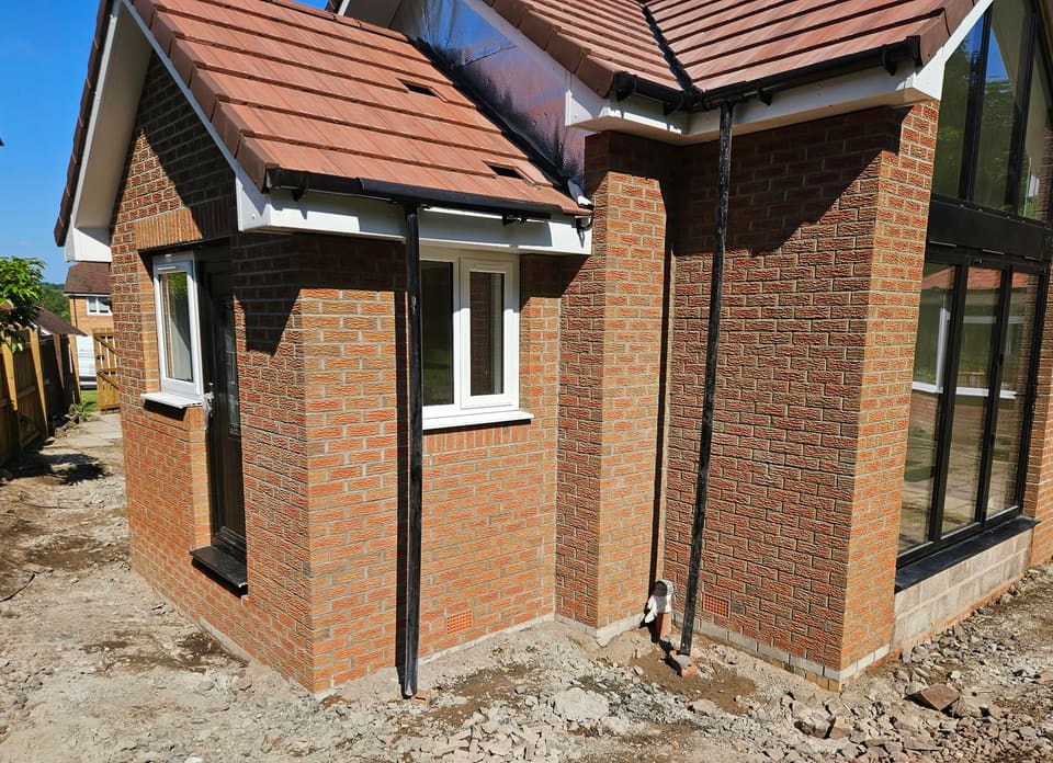 Corner of the brick extension with a black side door, white windows, two black downpipes and a brick pier beside the glazed gable.