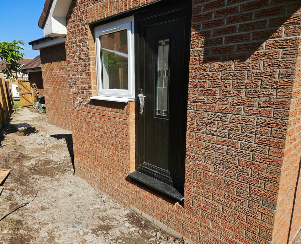Black side door and white window set in textured red brick under a soldier course, with a side passage running back to a timber gate.