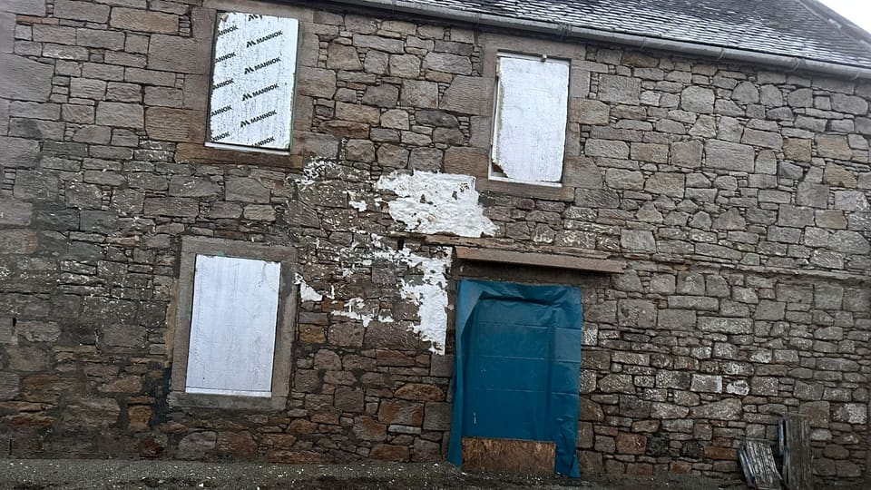 Two-storey sandstone rubble wall stripped of paint, windows boarded and doorway sheeted in blue tarpaulin.
