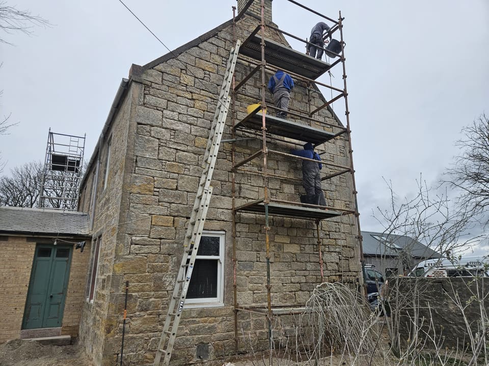 Three workers seen from behind on a three-lift tube scaffold against a sandstone gable, one hauling a tub on a rope. Ladder to the left.