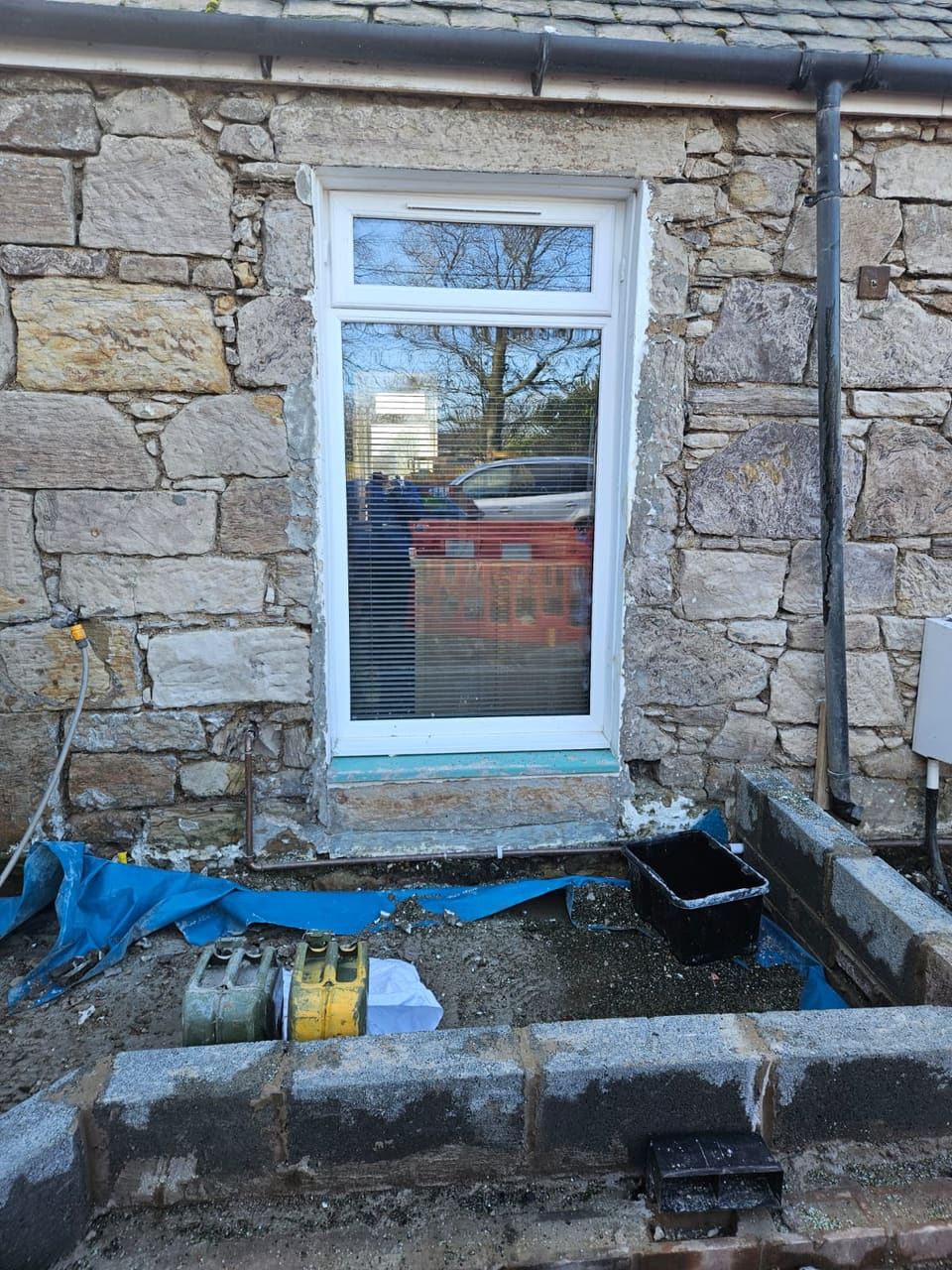 White window set straight into a rubble stone wall with no dressed margin, the ground below dug out and held back with blockwork.