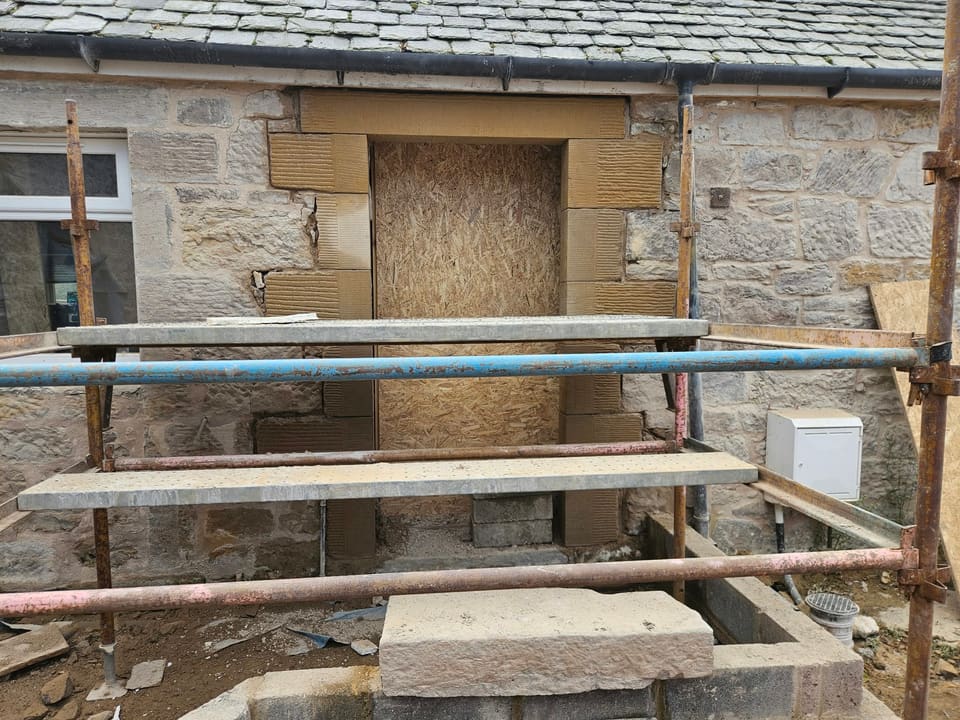 Front doorway boarded with OSB behind a scaffold, new sandstone rybats of alternating long and short stones under a new lintel, a large stone step in front.