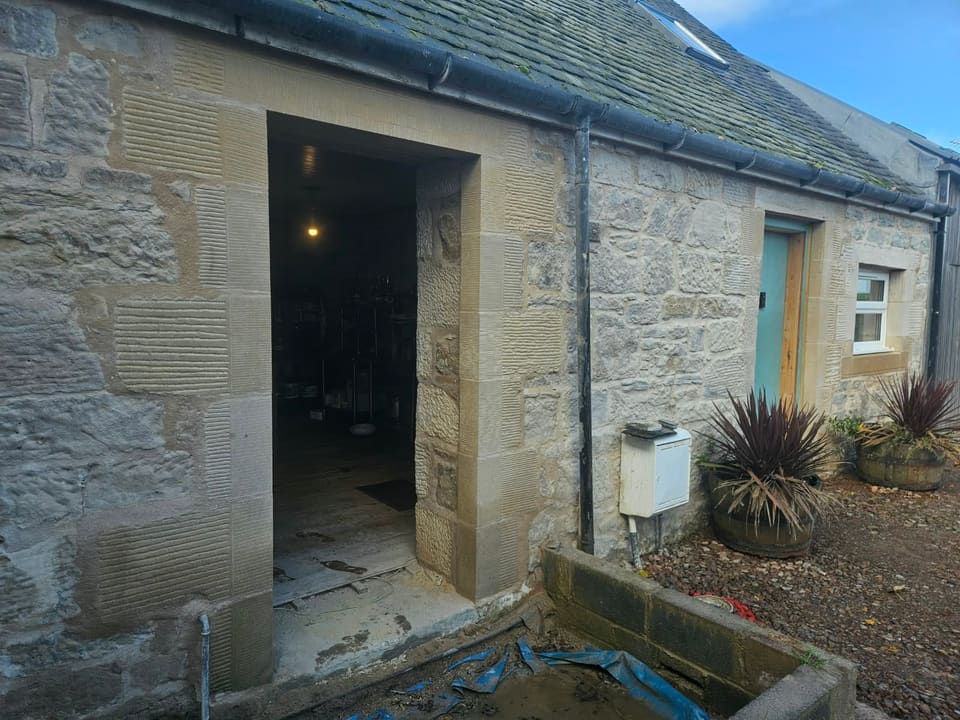 Front of the low stone building: the new sandstone doorway on the left, a downpipe and meter box, then the blue-green door in its dressed stone.