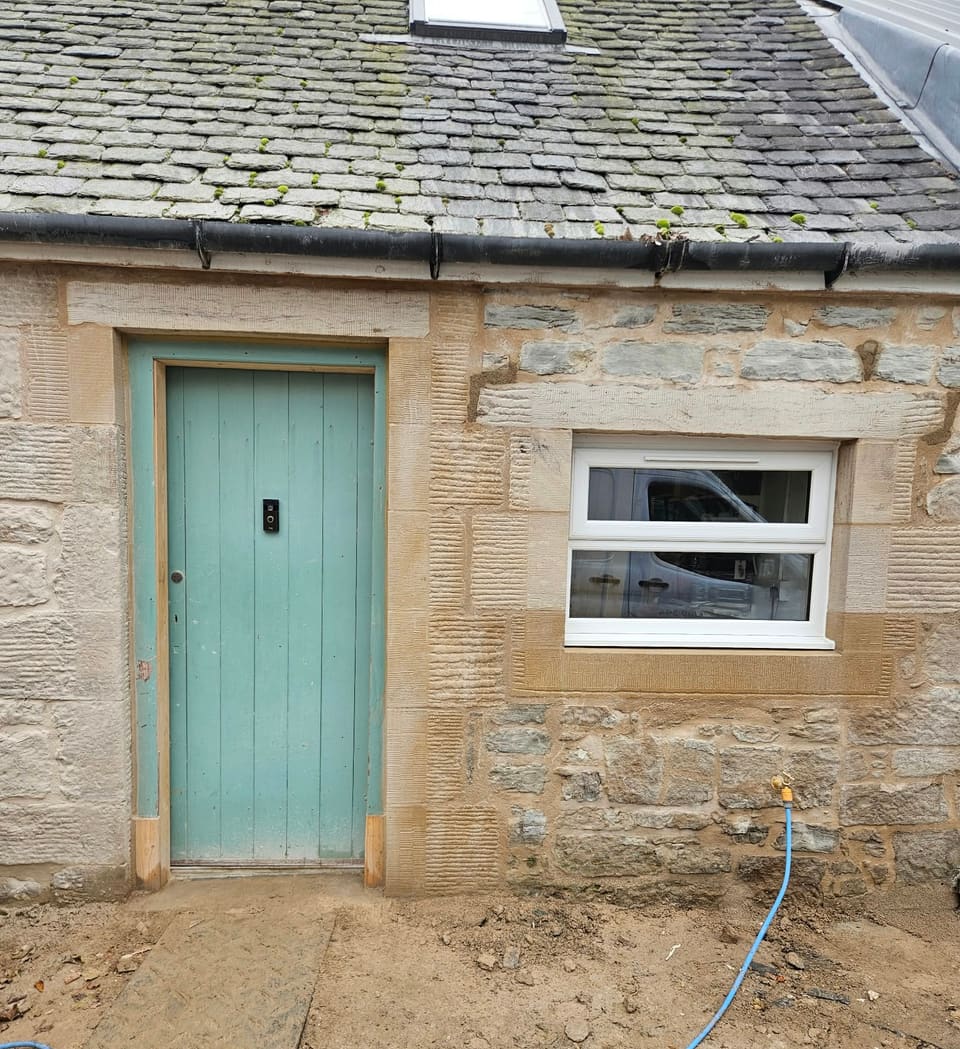 Blue-green boarded door and a small white window, both framed in dressed buff sandstone.