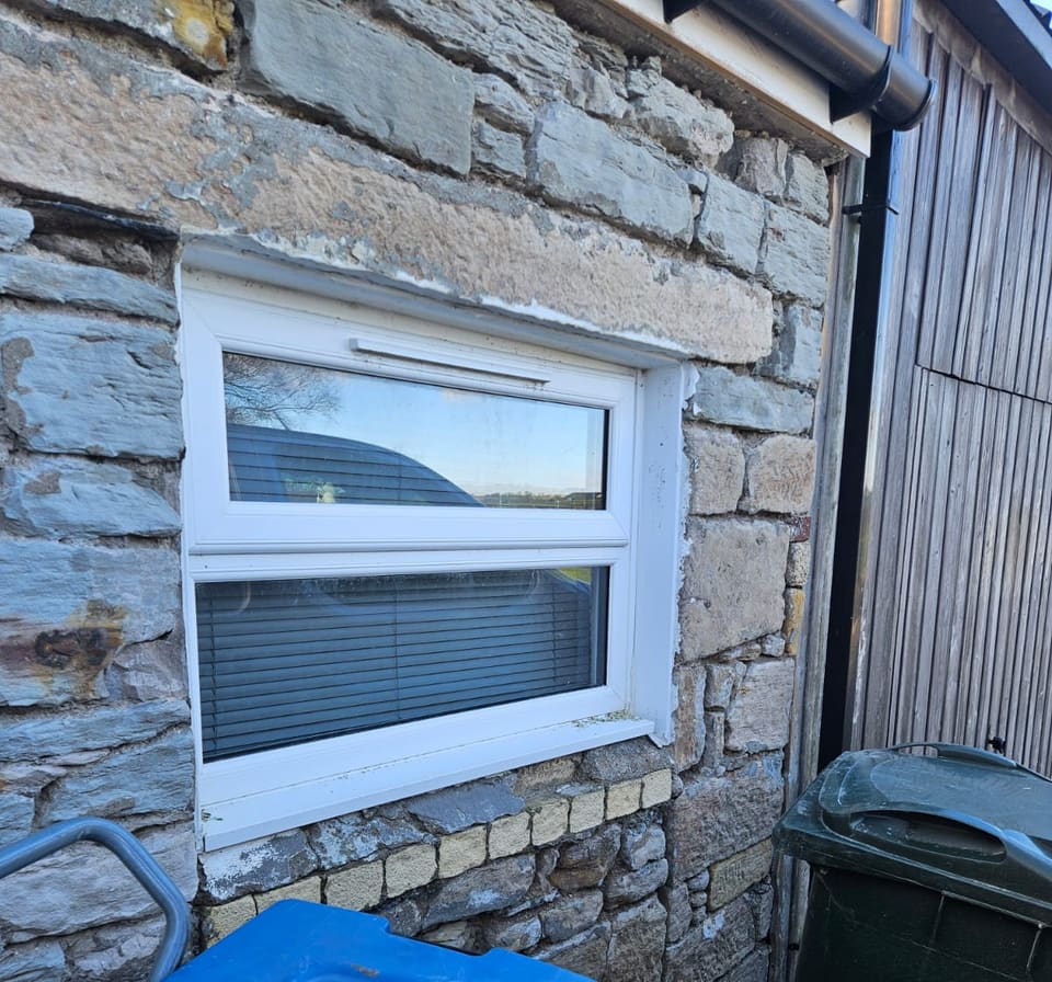 Small white window in a rubble wall beside a timber-clad building, with no dressed stone round it.