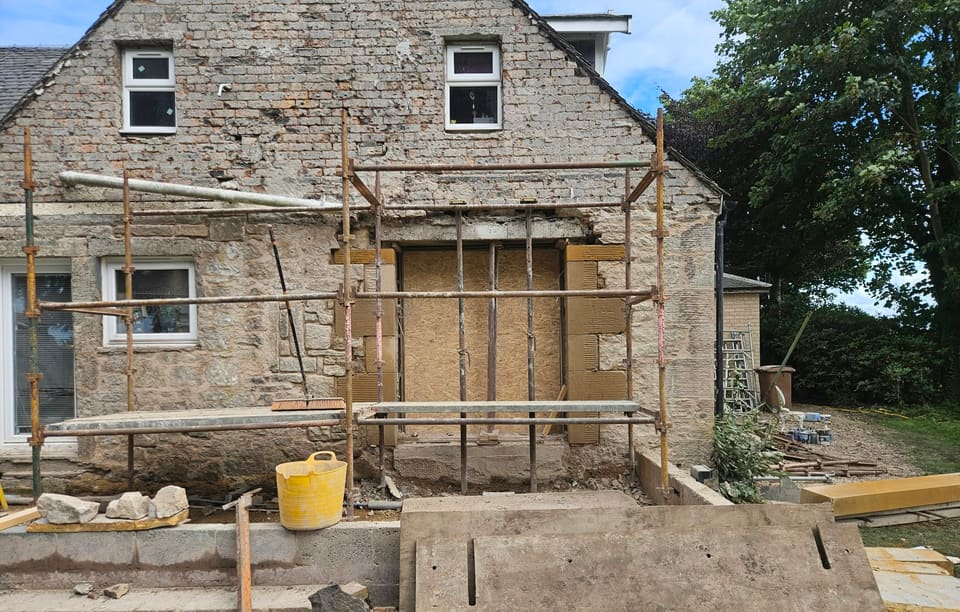 Two-storey stone gable with a scaffold round a wide ground-floor opening boarded with OSB, new sandstone rybats either side.