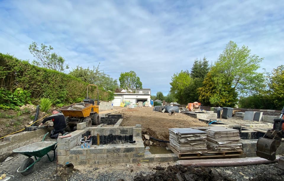 Dug-out garden with part-built blockwork walls coated in black, a dumper, pallets of paving slabs and a house with garages at the far end.