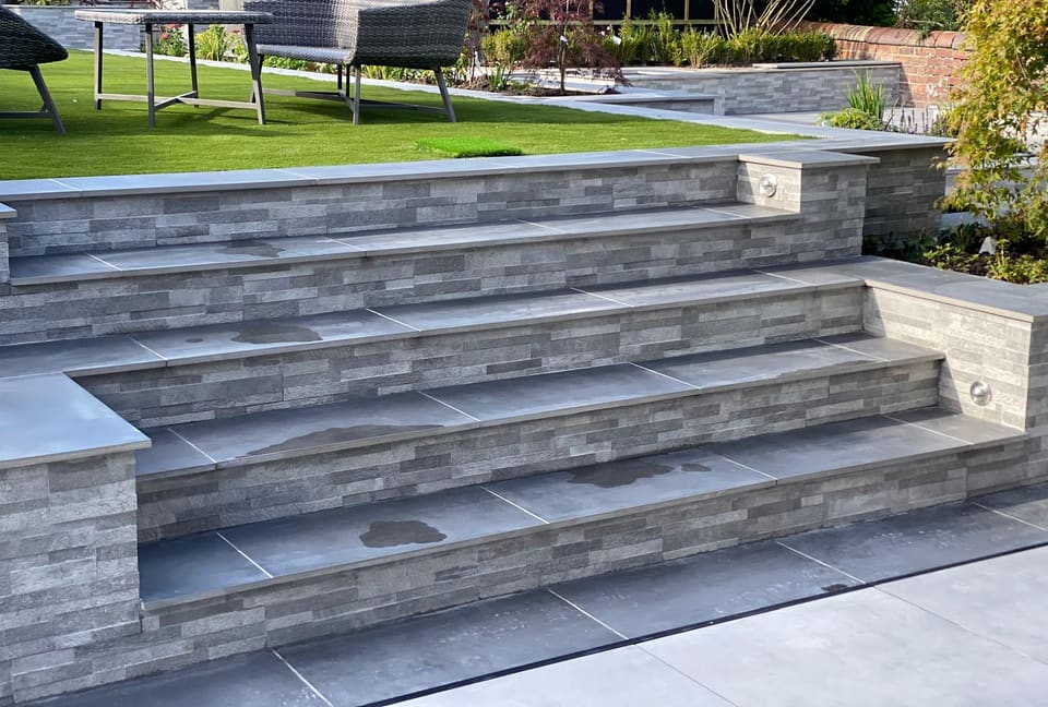 Wide flight of tiled steps with grey strip-clad risers rising to a lawn, flanked by clad walls with flat copings and two recessed lights.