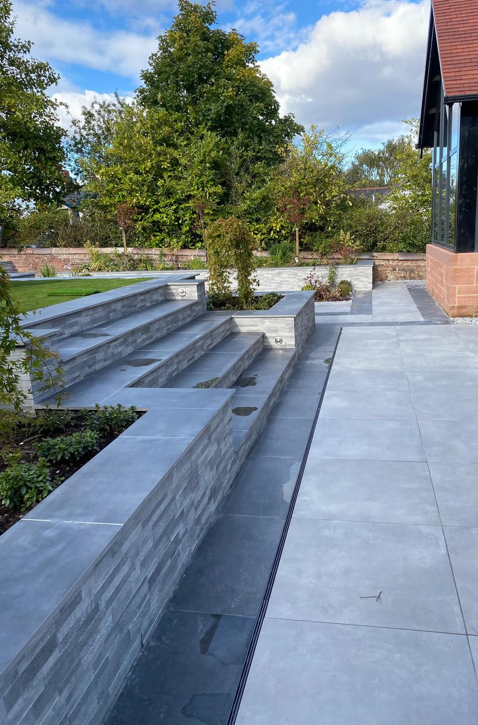 Side view along a planter coping and the steps towards the house, with a linear drain and a darker band running through the grey paving.