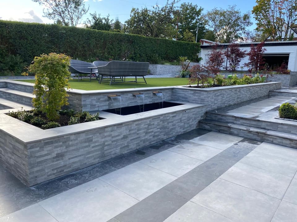 Three lit water spouts in a grey clad retaining wall pour into a raised dark pool, with planters either side and steps to the right.