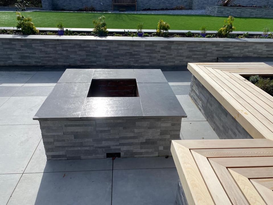 Square fire pit clad in the same grey strip cladding with a dark slab top, on the patio beside a clad planter wall.