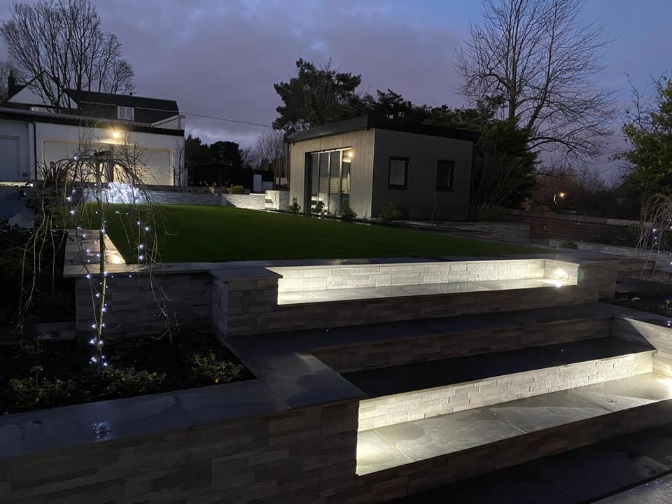 Dusk view of the lit steps between planters, with the lawn and a garden room behind.