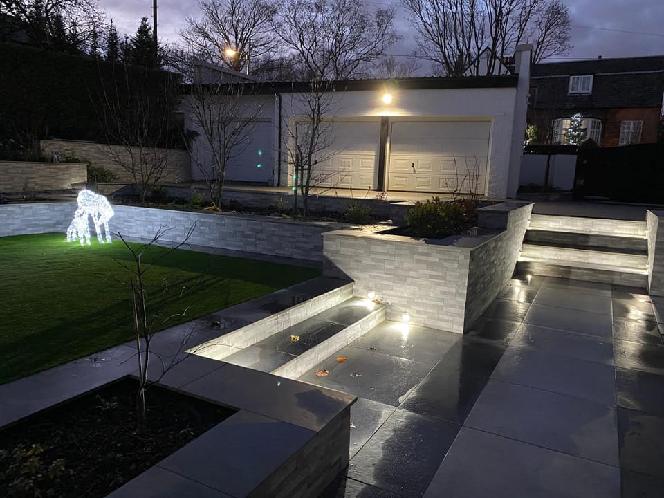 Dusk view of lit steps and a clad wall with small round wall lights, a white garage block behind.