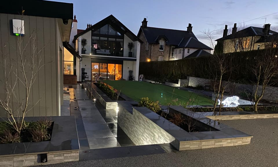 Dusk view along lit clad walls and dark paving towards the house, the lawn beyond.