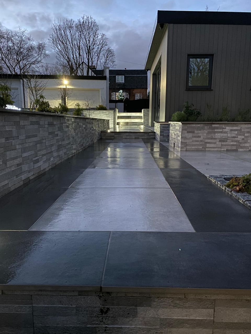 Dark paved path running beside a clad wall at dusk towards a lit garage.