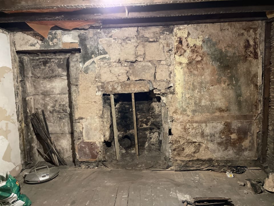 Bare rubble stone wall with patches of old plaster, an old fireplace opening with its lintel propped on two timbers, and a recess to the left.