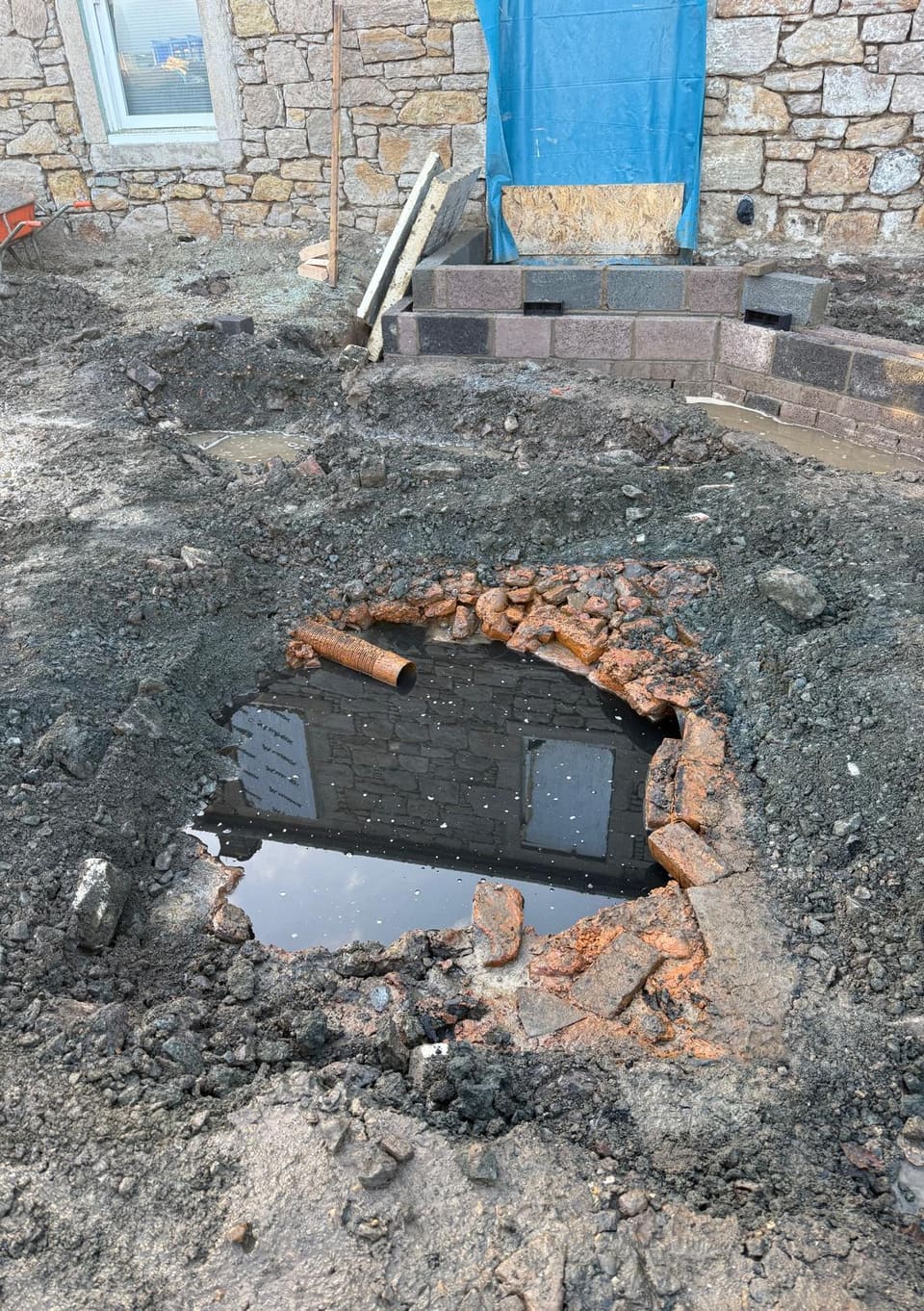 Ragged hole in dug ground in front of a stone house, ringed with broken red brick and full of dark water reflecting the house wall.