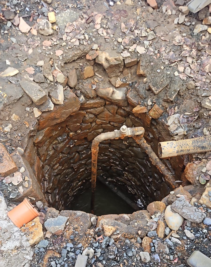 Round shaft lined with rust-stained rubble stone, an old rusty pipe and valve hanging inside, water below, loose rubble round the rim.