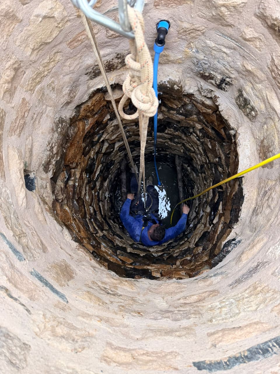 Looking down the well at a worker in blue on ropes at the bottom of the shaft, fixing a blue pipe to the dark original lining.