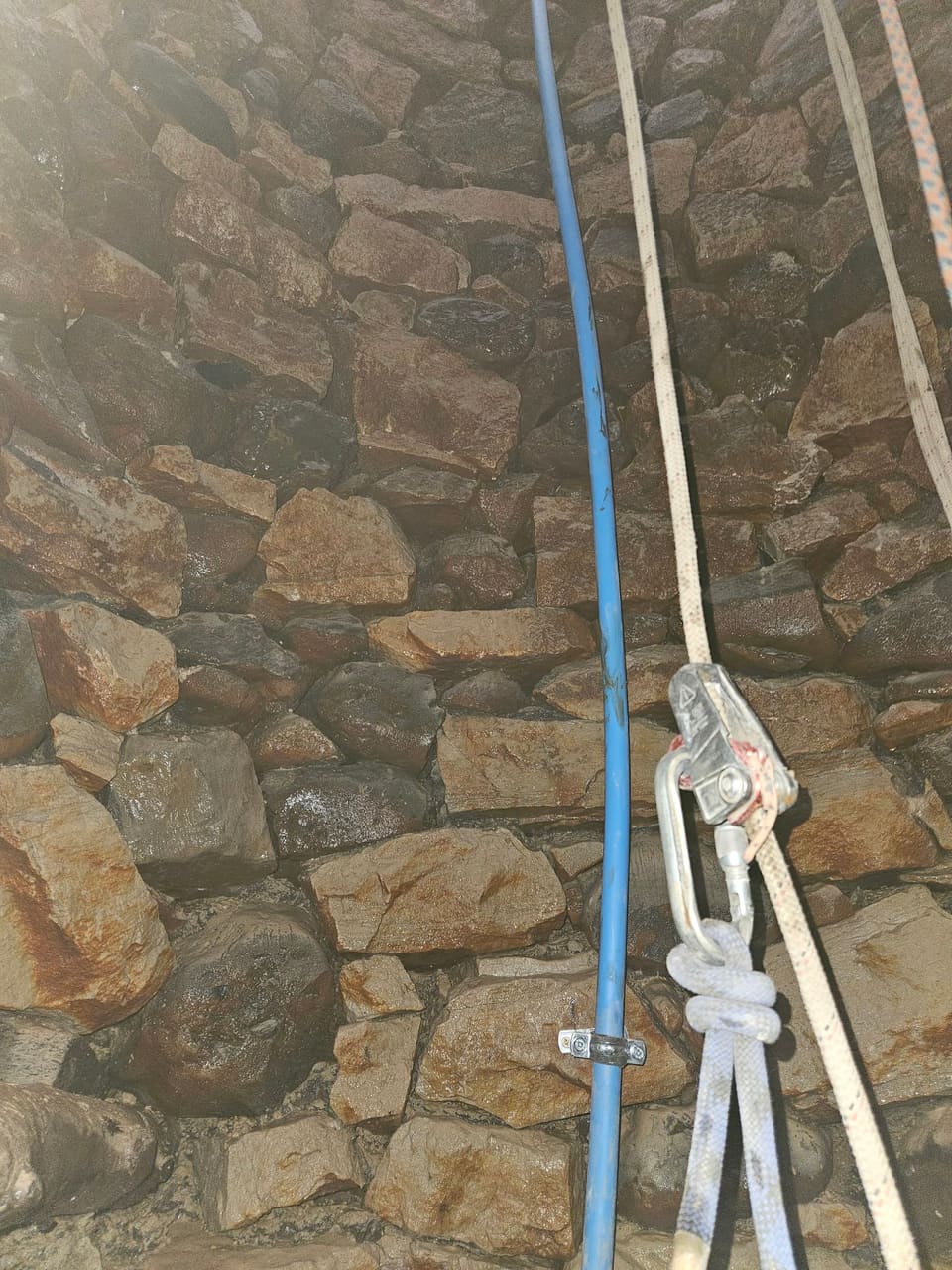 Blue pipe clipped to the original rubble lining of the shaft, beside the access ropes and a rope grab on a karabiner.