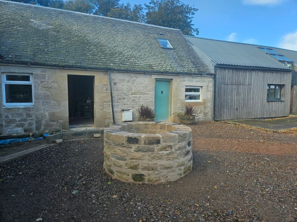 Finished round stone well head in a gravelled garden, in front of a low sandstone building with a slate roof and a timber-clad building to the right.