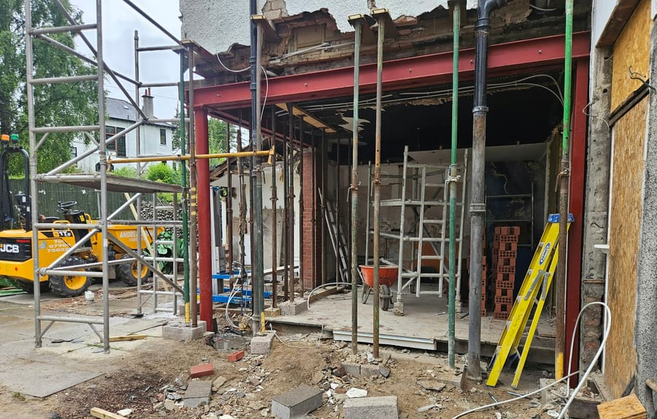 Back wall of a house opened up on a red steel frame, the floor above held on adjustable props, scaffold towers either side.