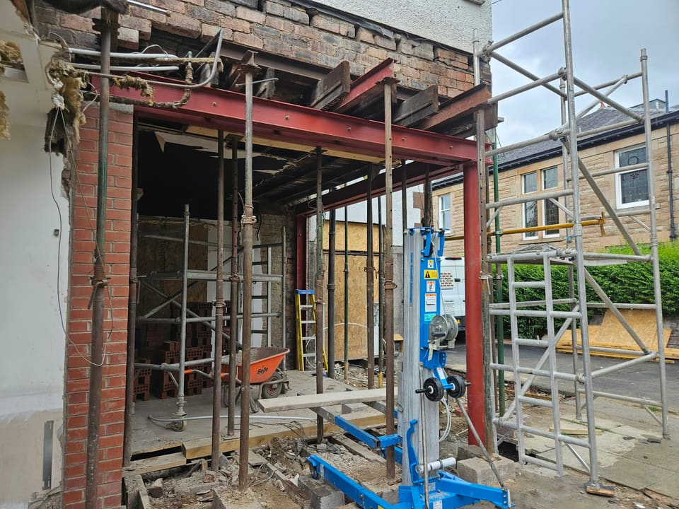 Red steel beam being raised into a wide new opening on a material lift, with props and a new brick pier.