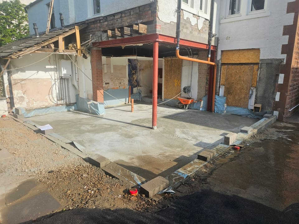 New concrete slab laid under a red steel goalpost frame across the opened-up back of a house.