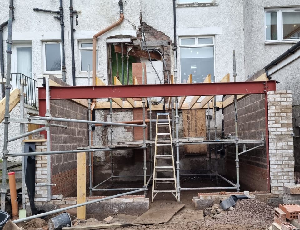 Extension shell with timber joists sitting on the red steel beam, a low scaffold inside and buff brick piers at both front corners.