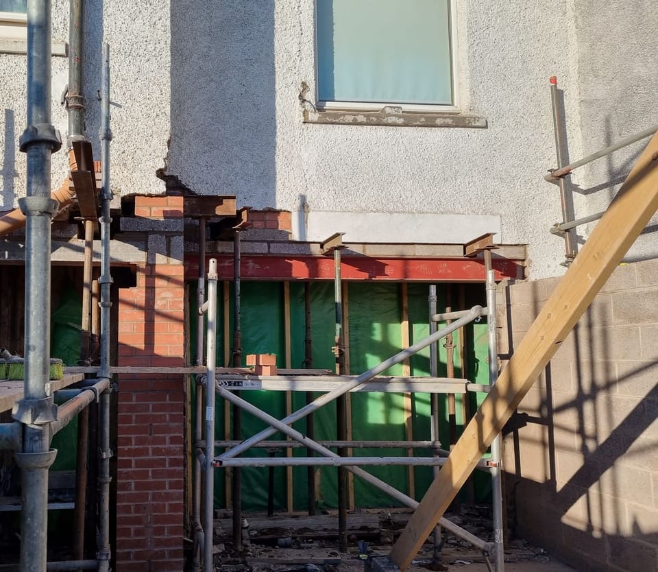 Rendered wall seen from a scaffold. A red steel beam spans a wide opening, with steel needles on props above and a brick pier to the left.