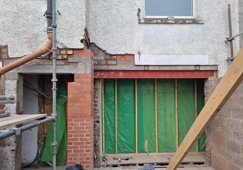 Rendered wall with a red steel beam over a wide opening screened in green sheeting. New red brick pier to the left. No props remain.